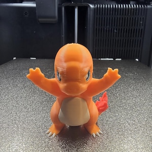 Peut inclure: Une figurine Charmander orange imprimée en 3D, avec les bras tendus, un ventre blanc et une queue rouge. La figurine a des yeux bleus et des griffes blanches. Elle est posée sur une surface grise texturée.