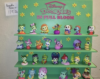 May include: A green display shelf featuring Disney Doorables figurines. The shelf is decorated with yellow flower accents and the text "Disney Doorables In Full Bloom". The figurines are colorful and depict various Disney characters.