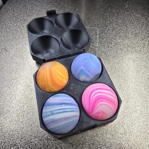 Op de afbeelding: Een zwarte kunststof tray met vier vakken met daarin vier bolvormige objecten met gemarmerde patronen. De bollen hebben verschillende kleurcombinaties met wervelingen in oranje en geel, lichtblauw en wit, blauw en paars, en roze en wit.