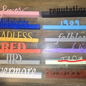 Pode incluir: Uma coleção de letreiros de prateleira coloridos, impressos em 3D, com várias palavras e frases. Os letreiros são rosa, preto, azul, azul claro, amarelo, laranja, cinza, roxo, verde e marrom. As palavras incluem "Lover", "Reputation", "Midnights", "1989", "Fearless", "Folklore", "Red", "Speak Now", "TPD", "I Love", "Evermore" e "The Life of a Showgirl".