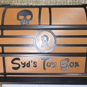 Puede incluir: Un cofre de juguete marrón y negro con dos estantes. El cofre de juguete tiene una calavera y las palabras "Syd's Toy Box". El cofre de juguete tiene un diseño de ojo de cerradura en el centro.