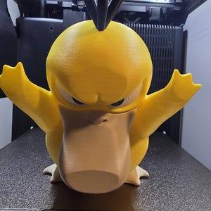 May include: A 3D-printed, yellow Psyduck figure with outstretched arms and an angry expression. The figure has a light brown beak and feet, and black hair. It is sitting on a black surface.