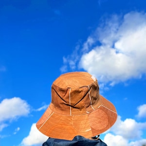Op de afbeelding: Een bruine vissershoed met witte verticale strepen en een brede rand. De hoed is gemaakt van een textuurstof. De hoed wordt gedragen door een persoon tegen een helderblauwe lucht met witte wolken.