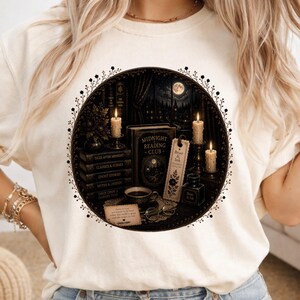 May include: A cream-coloured t-shirt featuring a circular graphic of a dark library scene with stacked books titled Tales After Midnight, Classics & Curses, Ghost Stories, Myths & Legends, and Once Upon a Dark Time, plus a Midnight Reading Club book.