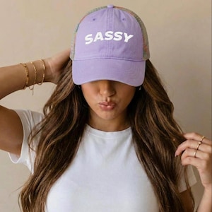 May include: A lavender baseball cap with the word "SASSY" in white block letters. The cap has a mesh back with a multicolored pattern. The person in the image is wearing a white t-shirt and gold bracelets.