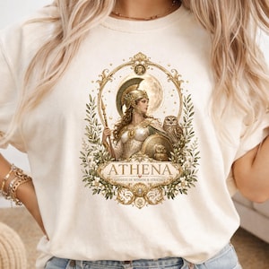 May include: A cream-colored t-shirt featuring a graphic print of the goddess Athena in golden armor with a spear, shield, and owl. The design is framed by an ornate gold border with olive branches and the text ATHENA GODDESS OF WISDOM & STRATEGY.