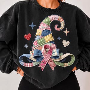 May include: A charcoal black crewneck sweatshirt featuring a large central graphic of a pointed hat constructed from various colorful patchwork fabric patterns. The design includes small star and heart shapes surrounding the hat, with a prominent bow.