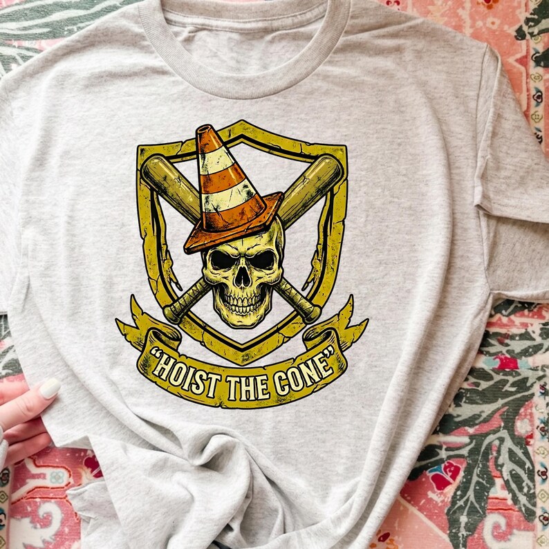 May include: Light gray t-shirt featuring a graphic design. The design includes a skull wearing a traffic cone, crossed baseball bats, and the text "HOIST THE CONE" on a banner. The graphic is in shades of yellow, orange, and brown.
