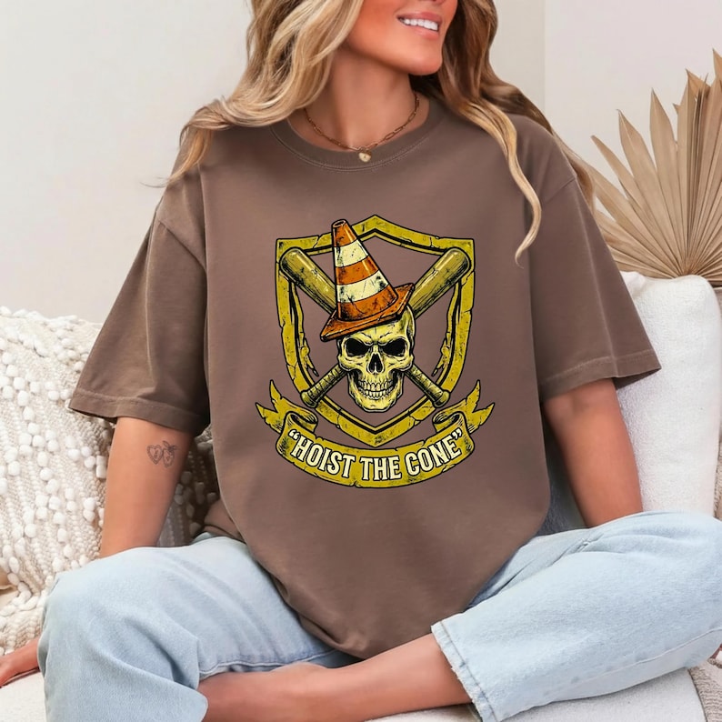May include: Brown t-shirt featuring a skull wearing a traffic cone hat, crossed baseball bats, and the text "HOIST THE CONE" in a banner. The design is in yellow and orange tones.