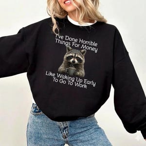 Puede incluir: Sudadera negra con un mapache sosteniendo una taza, con el texto: "He hecho cosas horribles por dinero, como levantarme temprano para ir a trabajar." El mapache es de color gris y marrón.