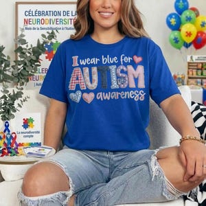 May include: A blue t-shirt with the text "I wear blue for AUTISM awareness" in a patchwork design. The shirt also features heart graphics. The image includes a framed sign with French text about neurodiversity and autism awareness.