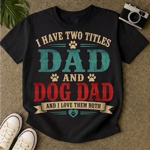 Può includere: T-shirt nera con la scritta "I HAVE TWO TITLES DAD AND DOG DAD AND I LOVE THEM BOTH". La parola "DAD" è turchese e "DOG DAD" è rossa. Sono presenti anche impronte di zampe e un cuore.