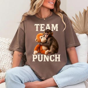 Może przedstawiać: Brązowa koszulka z napisem "TEAM PUNCH" w białych literach. Grafika przedstawia pluszową pomarańczową małpę obejmującą prawdziwą małpę. Koszulka ma luźny krój.