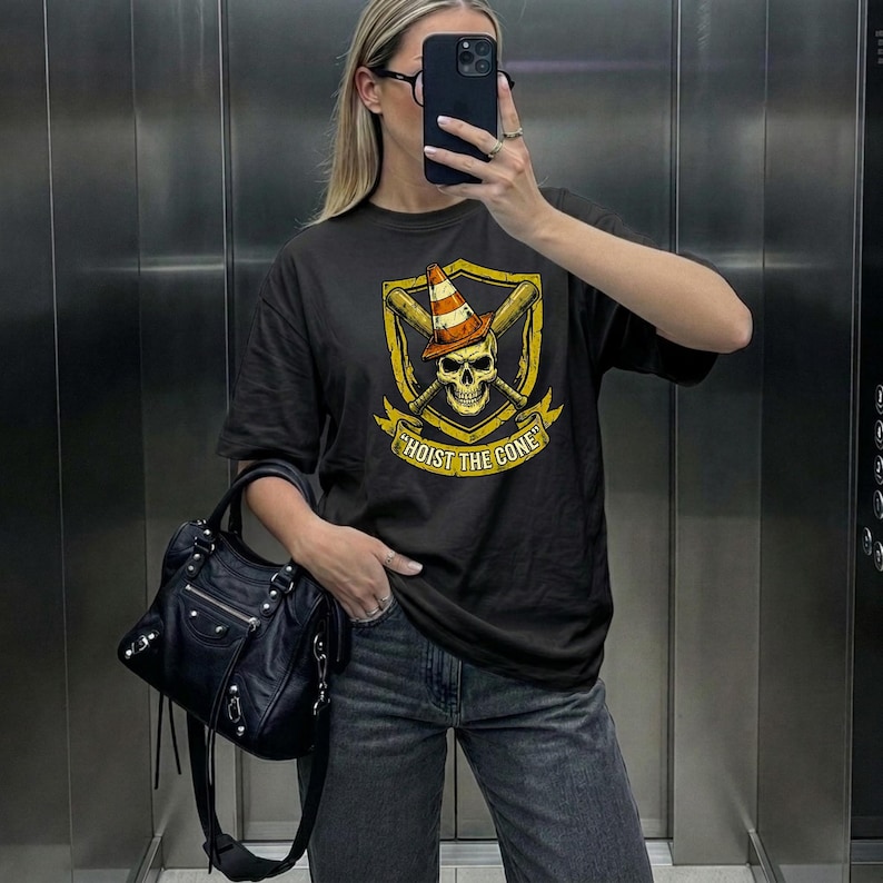 May include: Black t-shirt featuring a skull wearing a traffic cone, crossed baseball bats, and the text "HOIST THE CONE" in yellow. The design is set within a yellow shield. The shirt is paired with gray jeans and a black handbag.