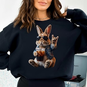 May include: Black sweatshirt featuring a cartoon rabbit wearing sunglasses and a denim jacket, holding a drink and making a rock-and-roll hand gesture. The rabbit is brown and white with long ears.