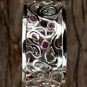 May include: A silver bangle bracelet with a delicate, intricate design featuring swirls and curves. The bracelet is adorned with small, pink gemstones.