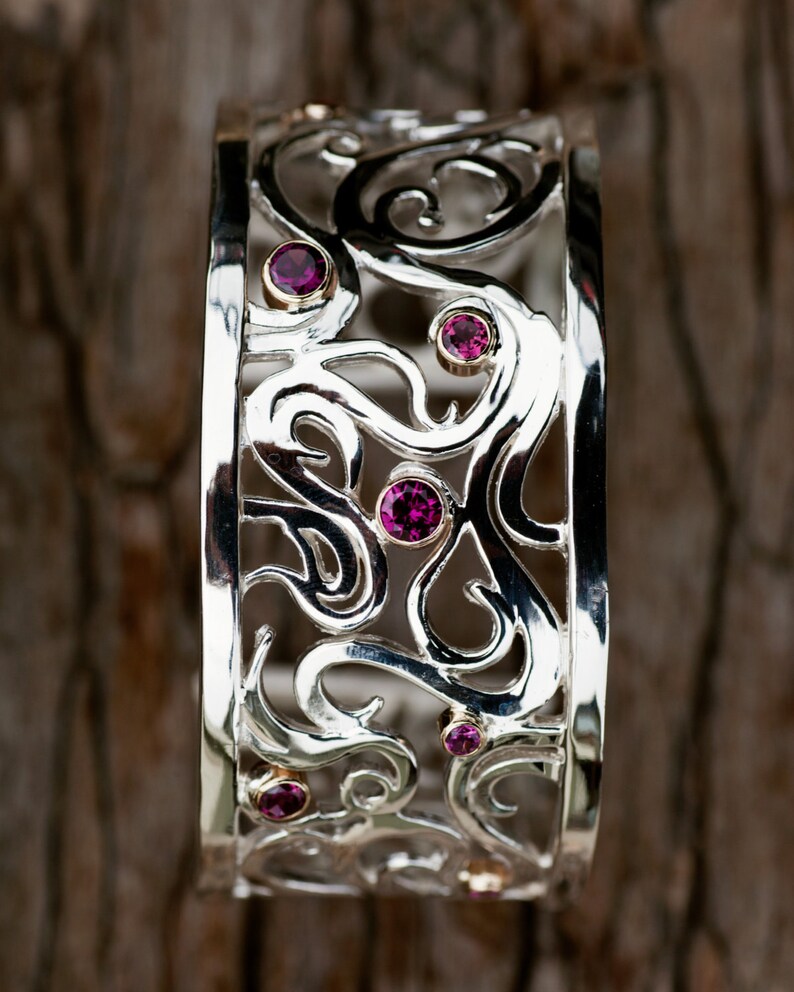 May include: A silver bangle bracelet with a delicate, intricate design featuring swirls and curves. The bracelet is adorned with small, pink gemstones.