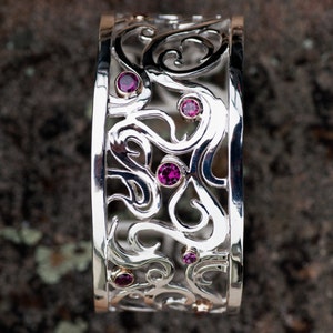 May include: A silver bangle bracelet with a delicate, intricate design featuring swirls and curves. The bracelet is adorned with five small, pink gemstones.