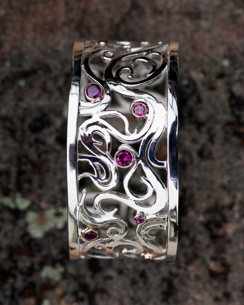 May include: A silver bangle bracelet with a delicate, intricate design featuring swirls and curves. The bracelet is adorned with five small, pink gemstones.