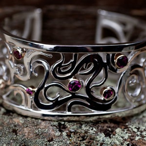 May include: A silver bangle bracelet with an intricate design featuring black swirls and purple gemstones.