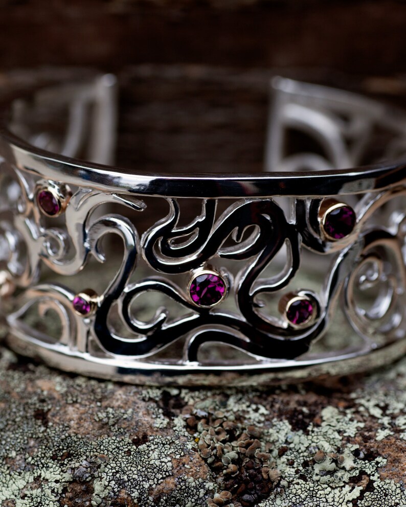 May include: A silver bangle bracelet with an intricate design featuring black swirls and purple gemstones.