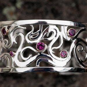 May include: A silver bracelet with a delicate, intricate design featuring swirls and curves. The bracelet is adorned with six small, pink gemstones.