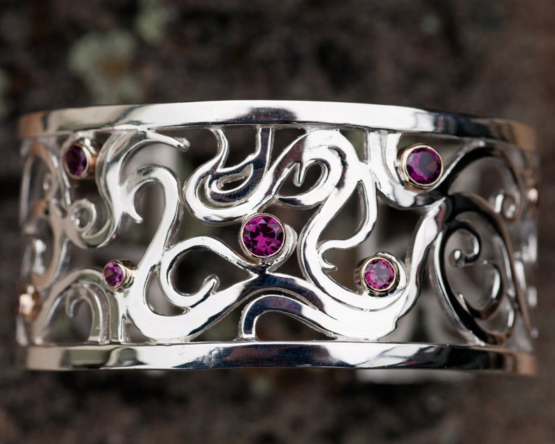 May include: A silver bracelet with a delicate, intricate design featuring swirls and curves. The bracelet is adorned with six small, pink gemstones.
