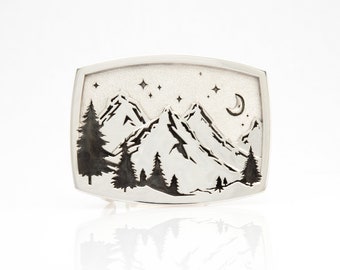 Puede incluir: Una hebilla de cinturón de plata con un diseño detallado de una cordillera de montañas bajo una luna creciente y estrellas.
