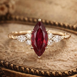 Puede incluir: Un anillo de oro con una piedra roja en forma de marquesa en el centro, flanqueada por piedras blancas redondas más pequeñas. El anillo tiene una banda texturizada y está sobre un fondo suave y neutro.