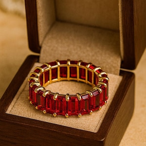 May include: A gold ring with a continuous band of rectangular, red gemstones. The ring is displayed inside a dark brown wooden jewelry box with a beige interior.