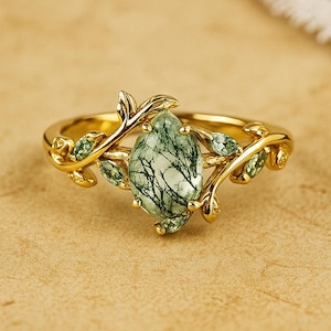 May include: A gold-colored ring featuring a large, oval-shaped green gemstone with a unique, tree-like pattern. The ring's band and setting are adorned with delicate, leaf-shaped details, creating an elegant, nature-inspired design.