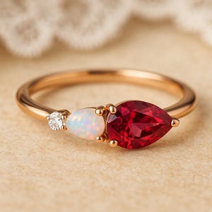 Puede incluir: Un anillo de oro rosa con tres piedras preciosas. Una pequeña gema redonda y transparente está engastada junto a un ópalo blanco en forma de pera. Una gema roja oscura más grande, en forma de pera, está en el lado opuesto del ópalo.
