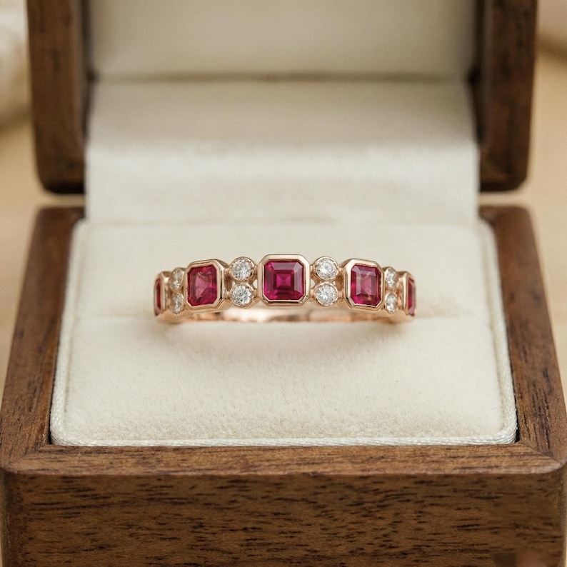 Pode incluir: Um anel de ouro rosa com pedras vermelhas quadradas e pequenas pedras claras redondas. O anel &eacute; exibido dentro de uma caixa de joias de madeira com um interior creme.