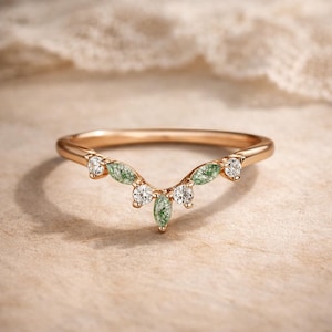 Peut inclure: Bague en or rose avec un design incurvé. La bague présente des pierres précieuses rondes et transparentes et des pierres vertes en forme de feuilles. La bague est posée sur un fond neutre, mettant en valeur les détails.