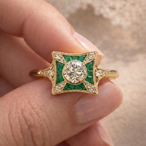 May include: A gold ring featuring a central round diamond, surrounded by emerald-colored stones and smaller diamonds. The ring has an Art Deco design with a square-shaped setting and a delicate band.