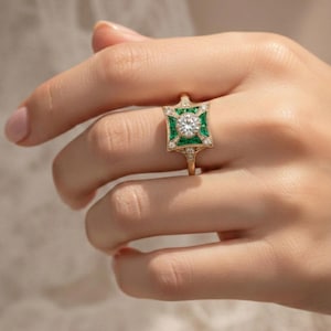 May include: A gold ring featuring a central round diamond surrounded by emerald-colored stones in a square design. The ring has a vintage, art deco style with small accent diamonds. The ring is held by a hand.