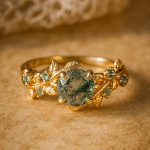 Op de afbeelding: Een gouden ring met een grote, ronde, groene edelsteen. De band van de ring is versierd met gedetailleerde bladeren en kleine edelsteenaccenten. De ring staat tegen een warme, neutrale achtergrond, wat het elegante ontwerp benadrukt.