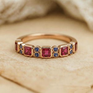 May include: Rose gold ring featuring square-cut red gemstones alternating with pairs of small blue gemstones. The ring has a continuous design with a polished finish, suitable for everyday wear or special occasions.