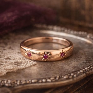 Peut inclure: Une bague en or ornée de trois motifs en forme d'éclat d'étoile, chacun serti d'une petite pierre précieuse rouge ronde. La bague est présentée sur un plateau en argent vintage, mettant en valeur son design élégant. La bande de la bague mesure environ 0,64 cm de large.