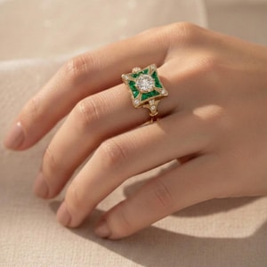 May include: A gold ring with a square emerald and diamond design. The ring features a central round diamond surrounded by green emeralds and smaller diamonds. The ring is on a person's finger.