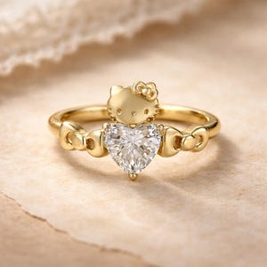 May include: A gold ring featuring a heart-shaped diamond and a Hello Kitty design. The ring has a gold band with bow-shaped accents. The Hello Kitty figure is at the top of the ring, with a bow on its head. The ring is set against a neutral background.