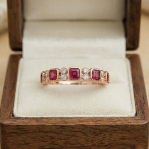 Pode incluir: Um anel de ouro rosa com altern&acirc;ncia de pedras vermelhas quadradas e pequenas pedras redondas e transparentes. O anel &eacute; exibido dentro de uma caixa de joias de madeira com interior creme.