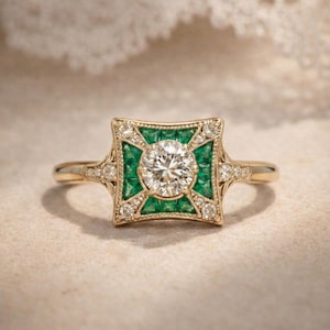 May include: A gold ring featuring a central round diamond, surrounded by a square frame of emeralds and smaller diamonds. The ring has a vintage, art deco style with intricate detailing and a polished finish.