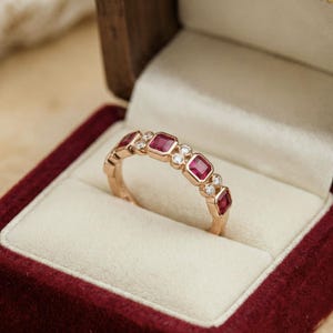 Pode incluir: Um anel de ouro com pedras vermelhas quadradas alternadas e pequenas pedras claras redondas. O anel &eacute; apresentado numa caixa de joias de veludo vermelho com interior creme. O anel &eacute; uma joia.