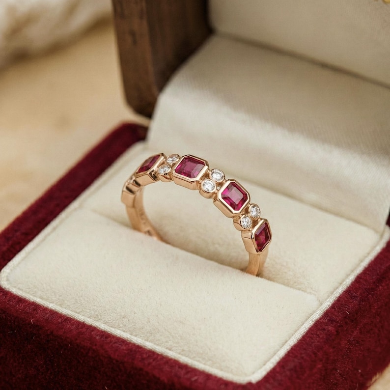 Pode incluir: Um anel de ouro com pedras vermelhas quadradas alternadas e pequenas pedras claras redondas. O anel &eacute; apresentado numa caixa de joias de veludo vermelho com interior creme. O anel &eacute; uma joia.