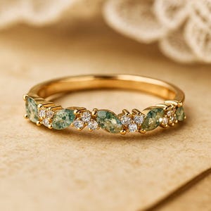 May include: A gold ring with a repeating pattern of green marquise-cut gemstones and small, clear gemstones. The ring is set on a gold band and is displayed on a neutral surface.