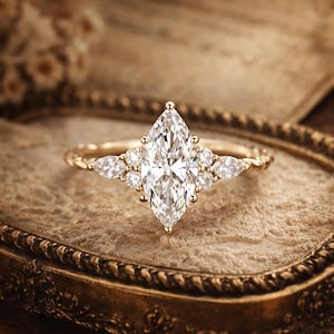 May include: A gold ring with a large, marquise-cut diamond as the centerpiece, flanked by smaller diamonds. The ring has a delicate, textured band and is displayed on a vintage-style, ornate tray.