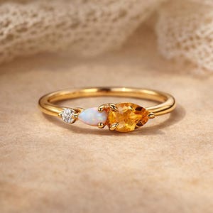 Könnte beinhalten: Ein goldener Ring mit drei Edelsteinen: einem kleinen Diamanten, einem birnenförmigen Citrin und einem ovalen Opal. Der Ring hat ein schmales Band. Der Citrin hat eine leuchtend orange Farbe.