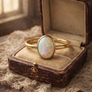 May include: A gold ring with an oval opal gemstone, displayed in a vintage brown jewelry box. The opal has iridescent colors. The ring is set on a beige velvet interior, with a lace doily in the background.