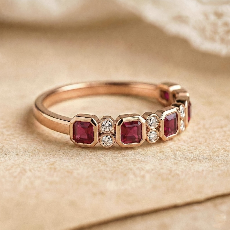 Pode incluir: Um anel de ouro rosa com uma s&eacute;rie de pedras preciosas vermelhas quadradas alternadas com pequenas pedras preciosas claras e redondas. O anel est&aacute; sobre um fundo neutro.